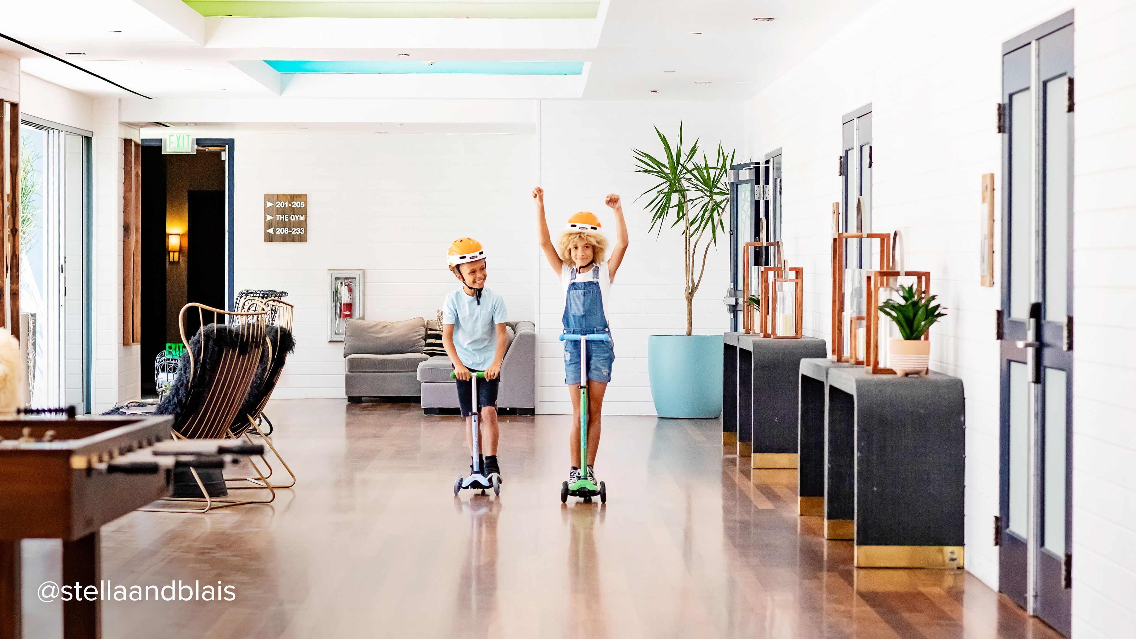 two kids with helmets riding micro scooteres inside a hotel lobby