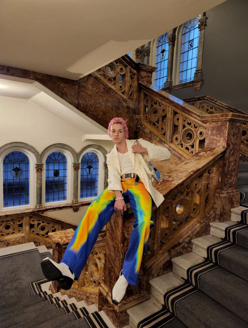 man dressed in rainbow hued trowsers and a white suite sitting on grand staircase bannister