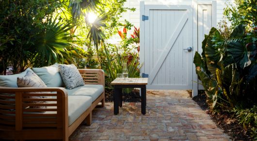 Outdoor paved pathway with lots of palms, a white gate, couch and table 