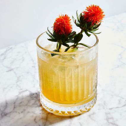 golden cocktail with red flower garnish on a white marble table